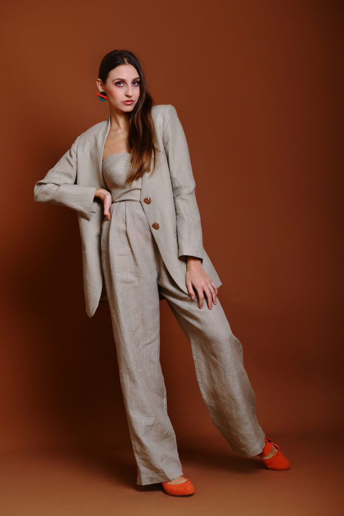 The girl wears a beige linen long blazer, a beige linen skirt and a red top, behind her is a beige background.