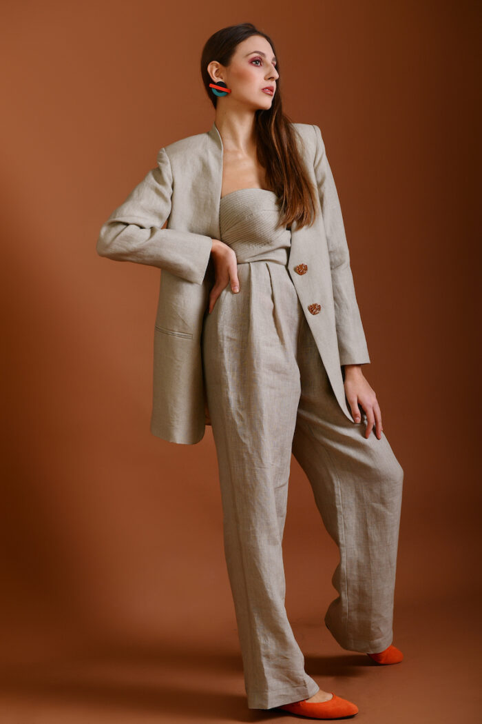 The girl wears a beige linen long blazer, a beige linen skirt and a red top, behind her is a beige background.