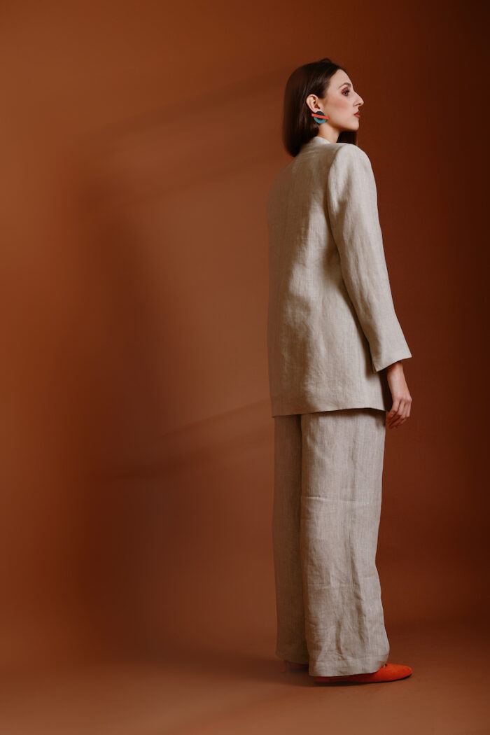 The girl wears a beige linen long blazer, a beige linen skirt and a red top, behind her is a beige background.