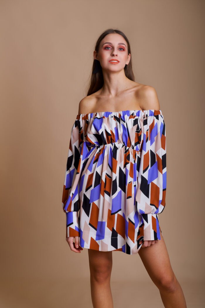 A girl wears a colorful off-the-shoulder silk MARIANA dress and stands in front of a beige background.