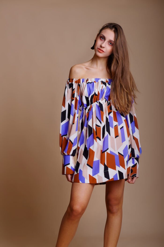 A girl wears a colorful off-the-shoulder silk MARIANA dress and stands in front of a beige background.