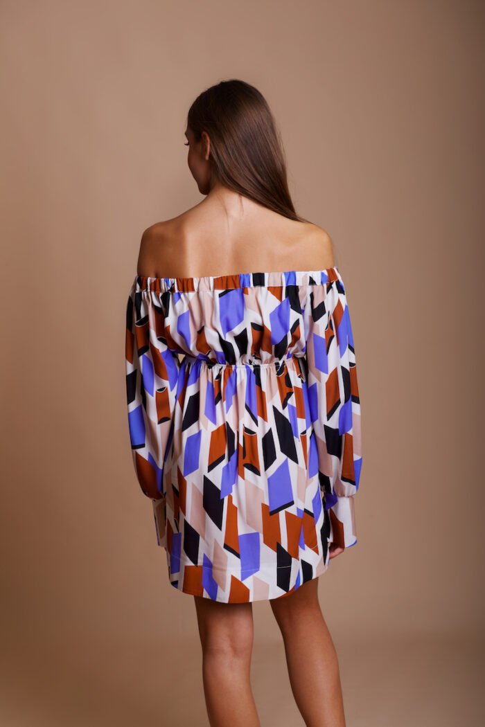 A girl wears a colorful off-the-shoulder silk MARIANA dress and stands in front of a beige background.