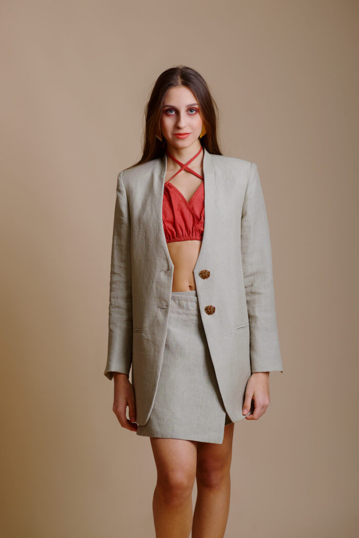 The girl wears a beige linen long blazer, a beige linen skirt and a red top, behind her is a beige background.