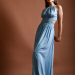 Devojka nosi dugačku DANICA haljina od svile sa golim ramenima. Stoji ispred pozadine tamno crvene boje.