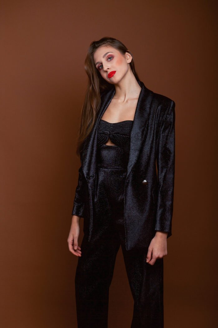 The girl wears a black LETITIA blazer with shiny sequins. He stands in front of a dark red background.