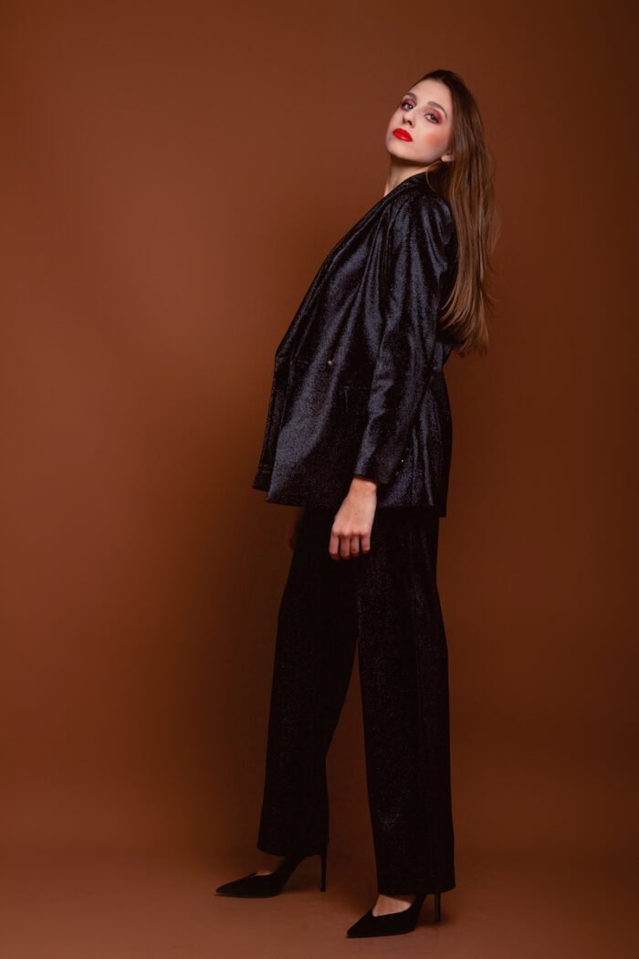 The girl wears a black LETITIA blazer with shiny sequins. He stands in front of a dark red background.