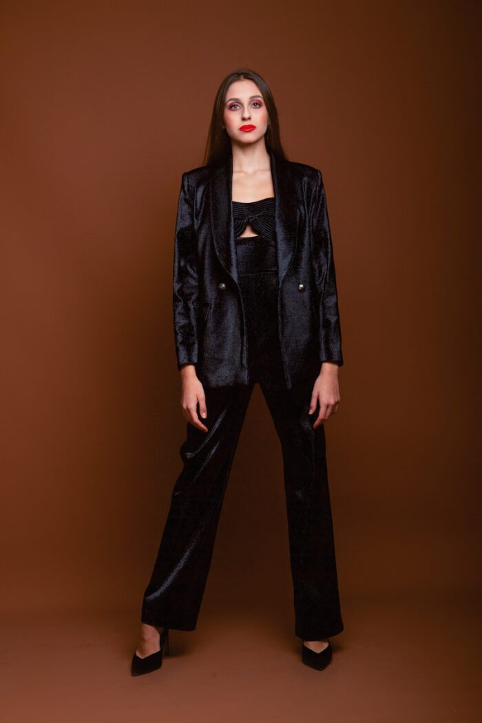 The girl wears a black LETITIA blazer with shiny sequins. He stands in front of a dark red background.