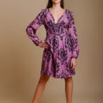 Devojka nosi kratku MARIA haljinu od dezenirane svile u ljubičastoj boji sa crnim cvetnim detaljima.