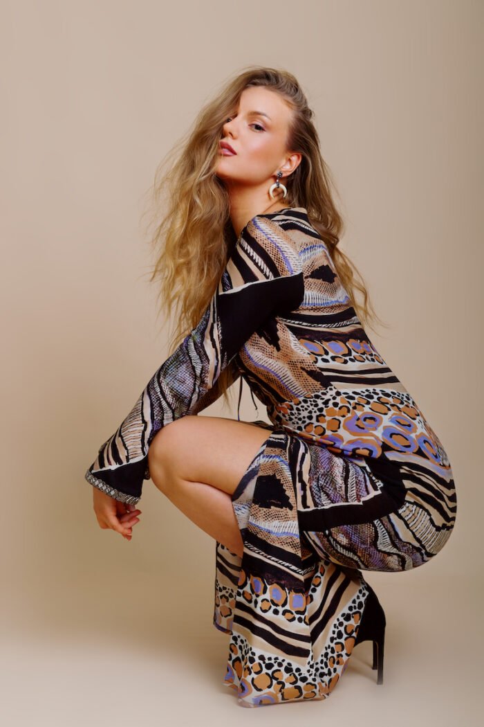 The girl wears a UNA dress made of patterned silk in an animal print, with lots of designer details. The girl wears black shoes with high heels and stands in front of a beige background.