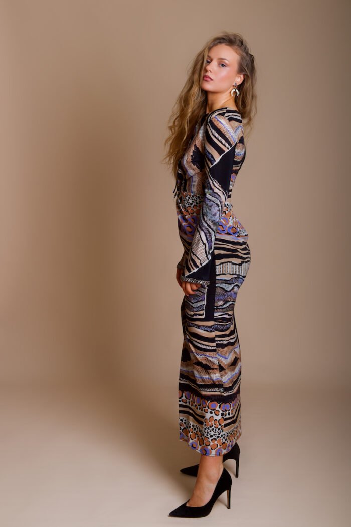 The girl wears the UNA dress made of patterned silk in an animal print, with lots of designer details. The girl wears black shoes with high heels and stands in front of a beige background.