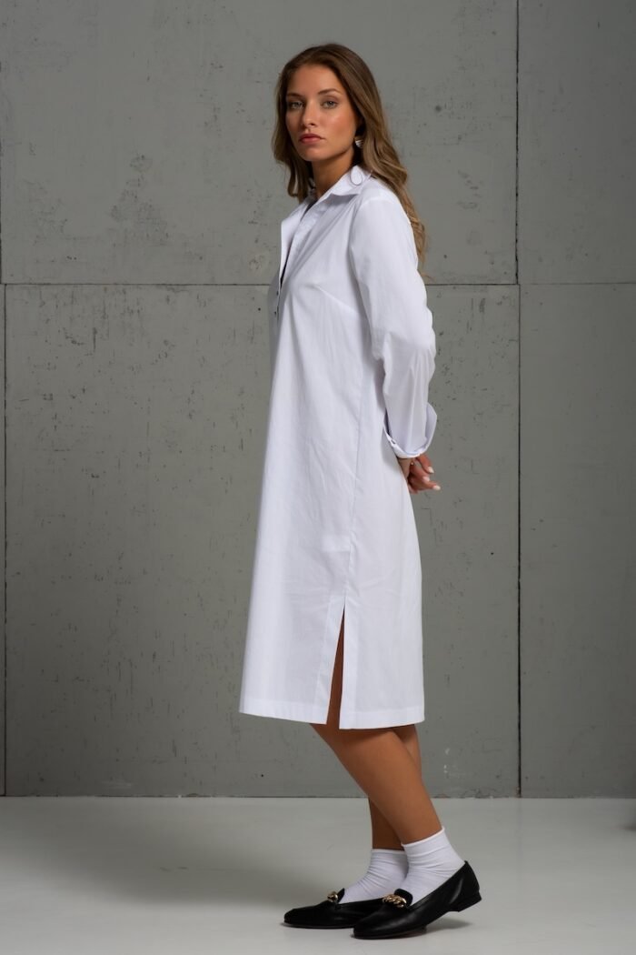 Brunette is wearing a white cotton shirt of midi length and standing in front of a gray background.