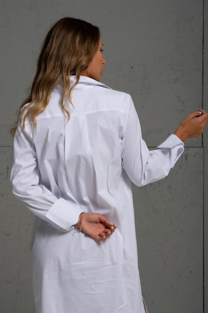 Brunette is wearing a white cotton shirt of midi length and standing in front of a gray background.