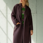 Plava devojka nosi ljubičasti kaput na vezivanje i stoji ispred sive pozadine.