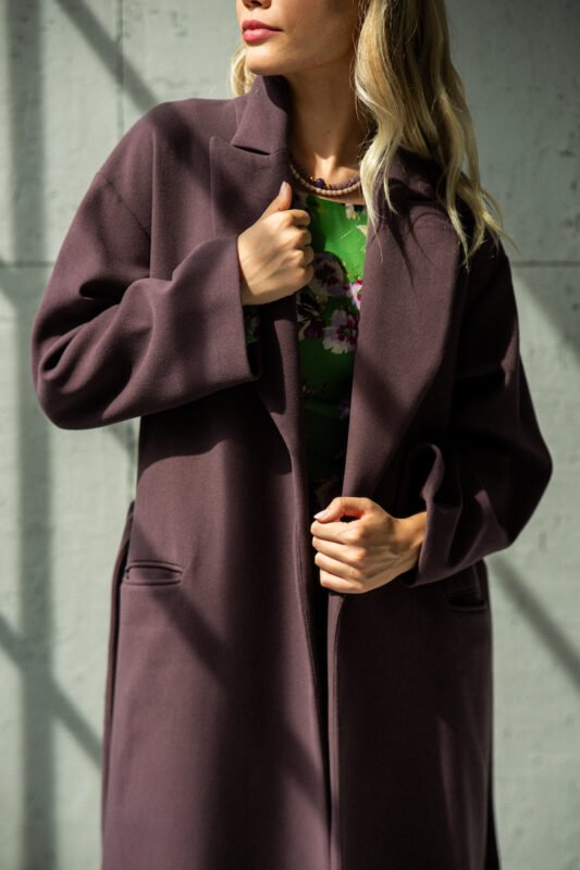 The blonde girl is wearing a purple tie-waist coat and standing in front of a gray background.