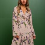 Brineta nosi kratku bež haljinu od svile sa floralnim printom i stoji ispred zelene pozadine.