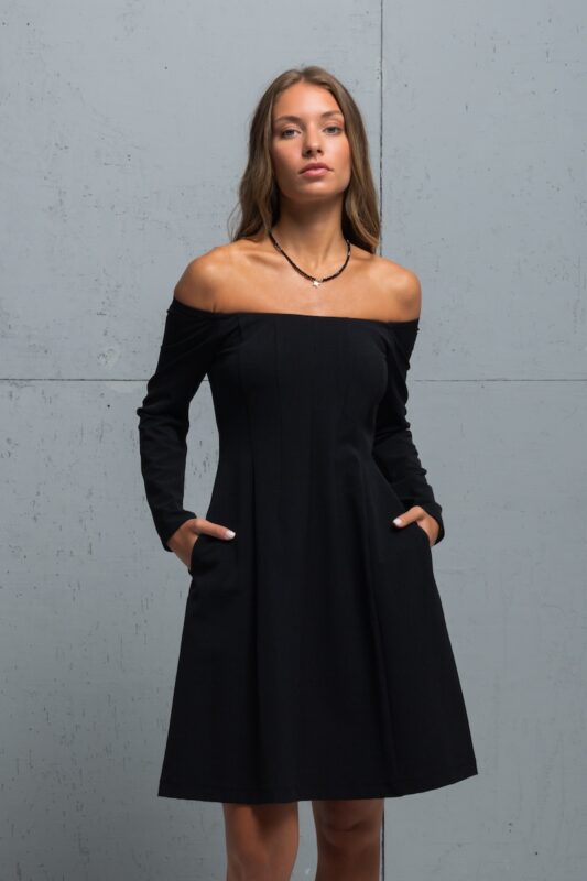 The brunette is wearing a little black dress with long sleeves and a Bardot neckline. She is standing in front of a gray wall.