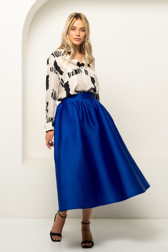 The blonde girl is wearing a blue BARBARA skirt and a white and black silk shirt and is standing in front of a white wall.