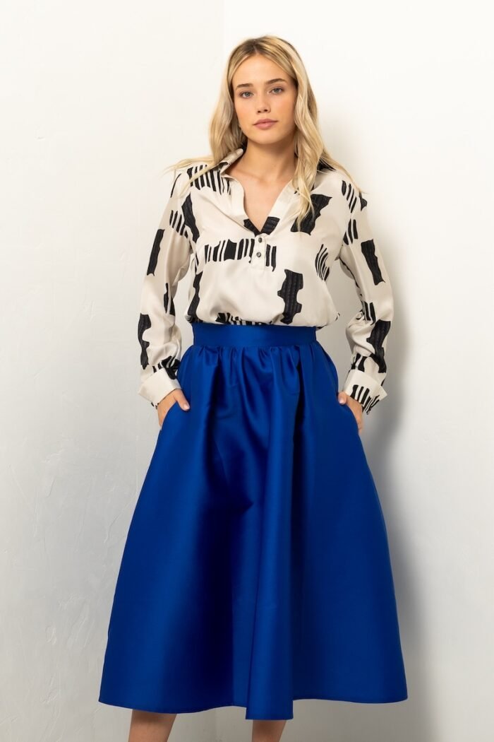 The blonde girl is wearing a blue BARBARA skirt and a white and black silk shirt and is standing in front of a white wall.