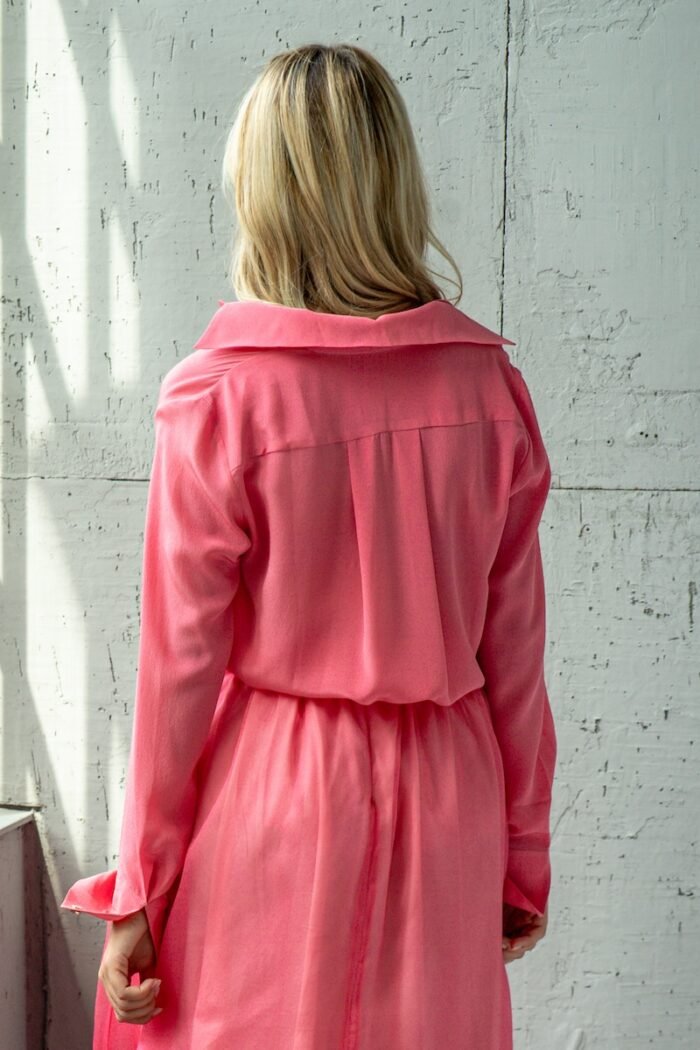 The blonde girl is wearing a light pink BELA silk shirt and standing in front of a gray wall.