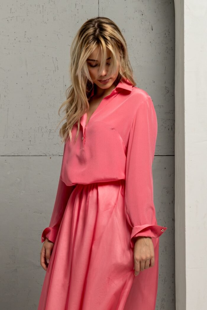 The blonde girl is wearing a light pink BELA silk shirt and standing in front of a gray wall.