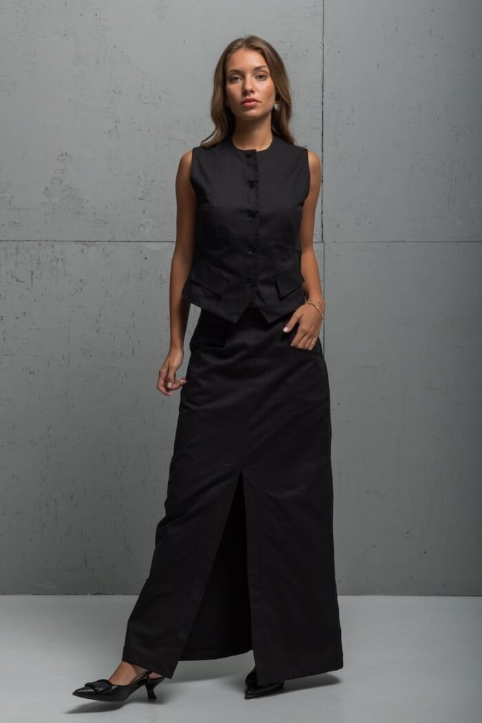 The girl is wearing a long skirt made of black twill with a slit at the front. She is standing in front of a gray wall.