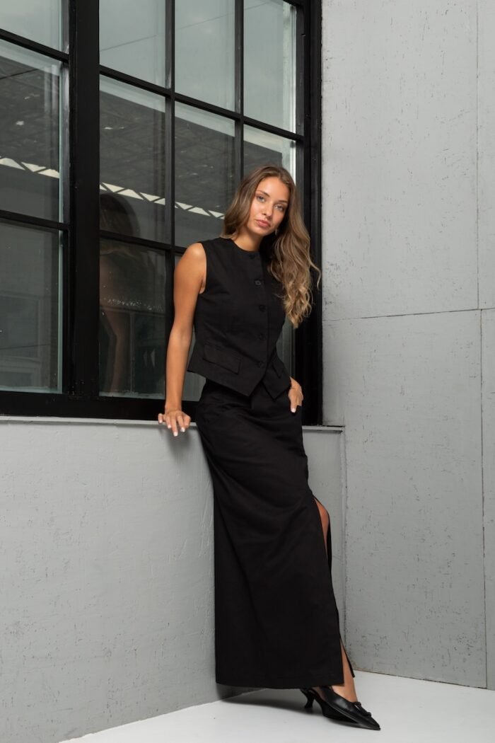 The girl is wearing a long skirt made of black twill with a slit at the front. She is standing in front of a gray wall.