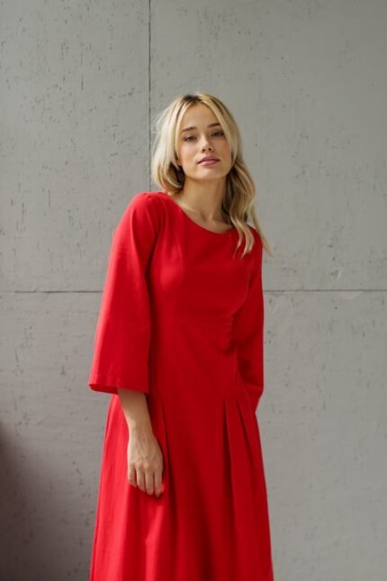 A blonde girl wears a red midi MAXIMA dress and stands in front of a gray wall.