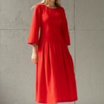 Plava devojka nosi crvenu midi MAXIMA haljinu i stoji ispred sivog zida.