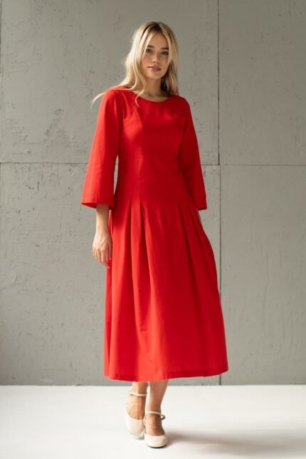 A blonde girl wears a red midi MAXIMA dress and stands in front of a gray wall.