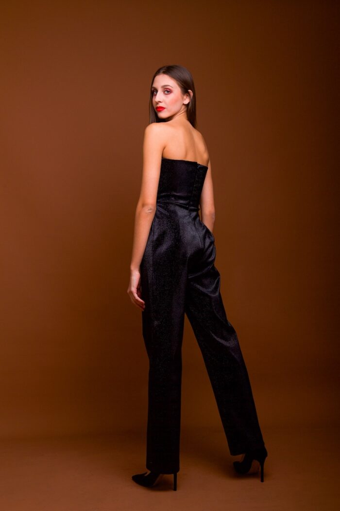 The girl wears a shimmering black jumpsuit with a prominent corset. She was photographed in front of a dark red background.