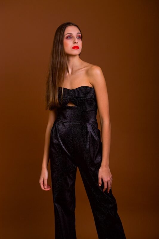 The girl wears a shimmering black jumpsuit with a prominent corset. She was photographed in front of a dark red background.