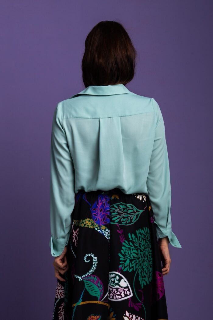 A woman stands in front of a purple background and wears a mint green blouse with a black patterned skirt.