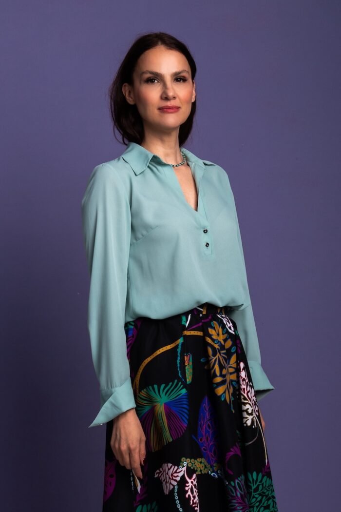 A woman stands in front of a purple background and wears a mint green blouse with a black patterned skirt.