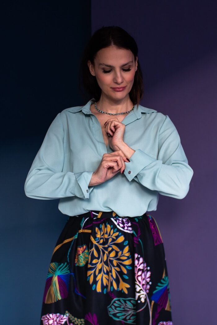 A woman stands in front of a purple background and wears a mint green blouse with a black patterned skirt.