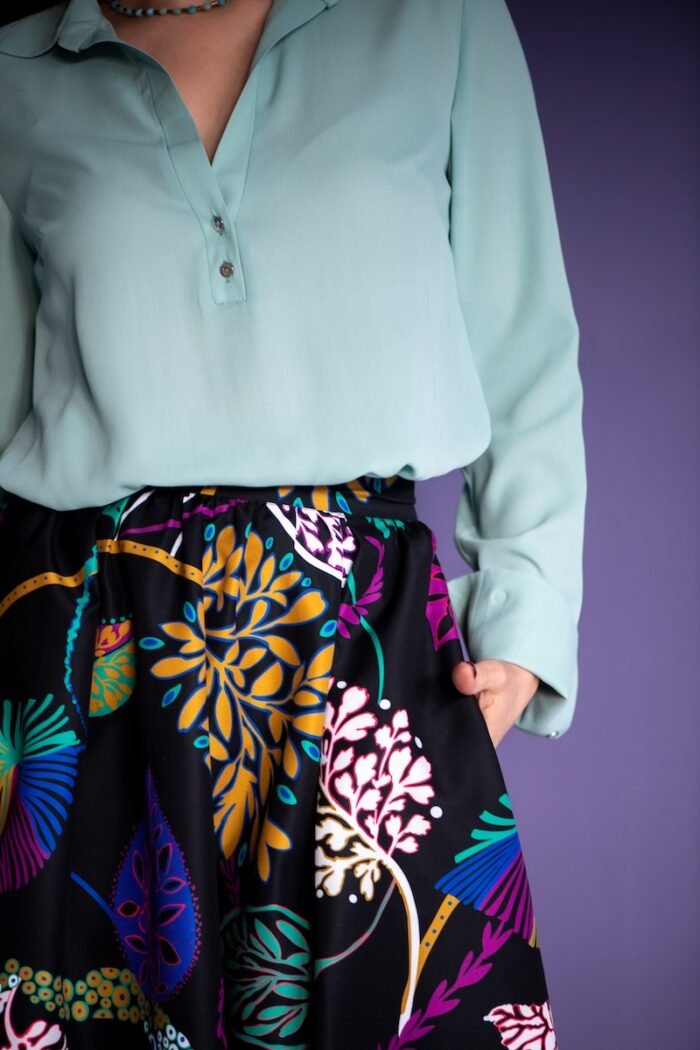 A woman stands in front of a purple background and wears a mint green blouse with a black patterned skirt.