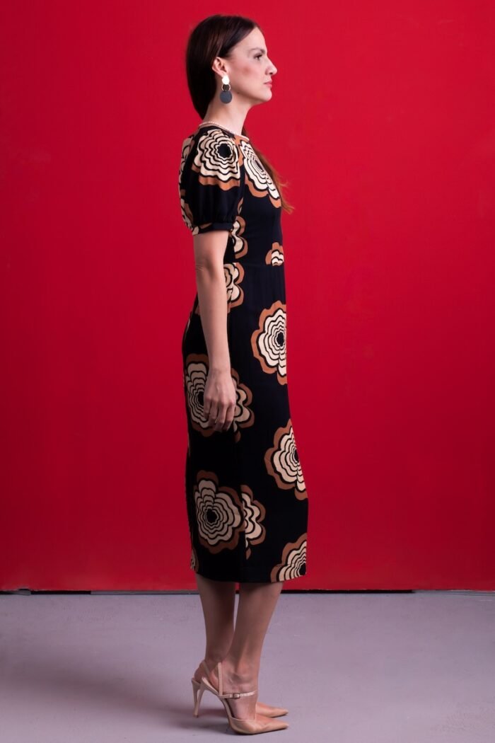 A woman wears a black dress with floral motifs, sitting on a chair in front of a red background.