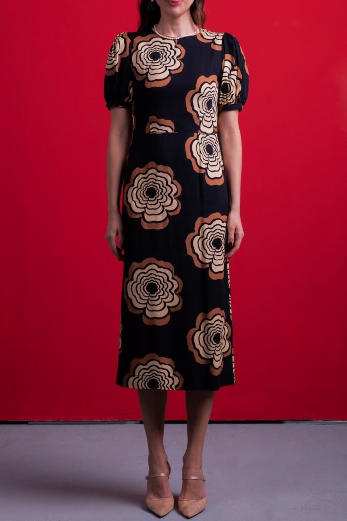 A woman wears a black dress with floral motifs, sitting on a chair in front of a red background.