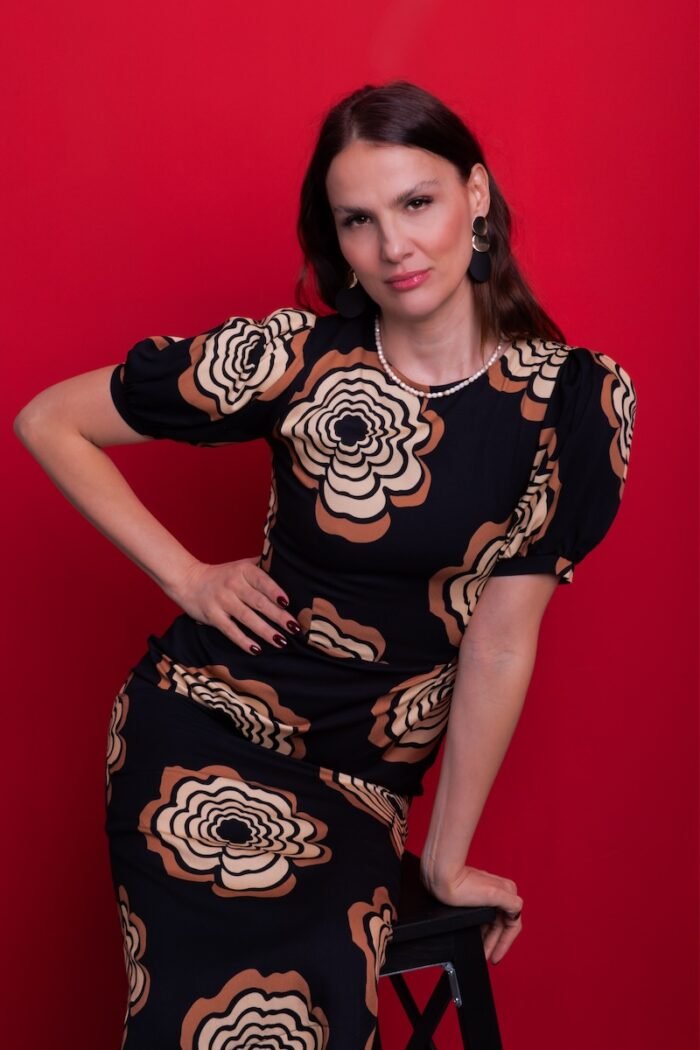 A woman wears a black dress with floral motifs, sitting on a chair in front of a red background.