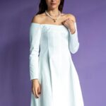 A woman wears a short white dress and stands in front of a purple wall.