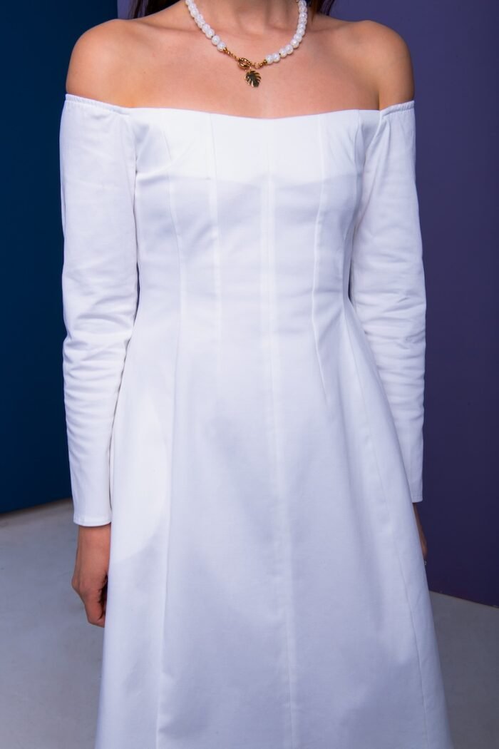 A woman wears a short white dress and stands in front of a purple wall.