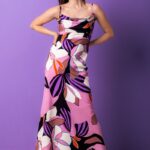 Žena nosi dugu, svilenu haljinu floralnog dezena. Stoji ispred ljubičaste pozadine.