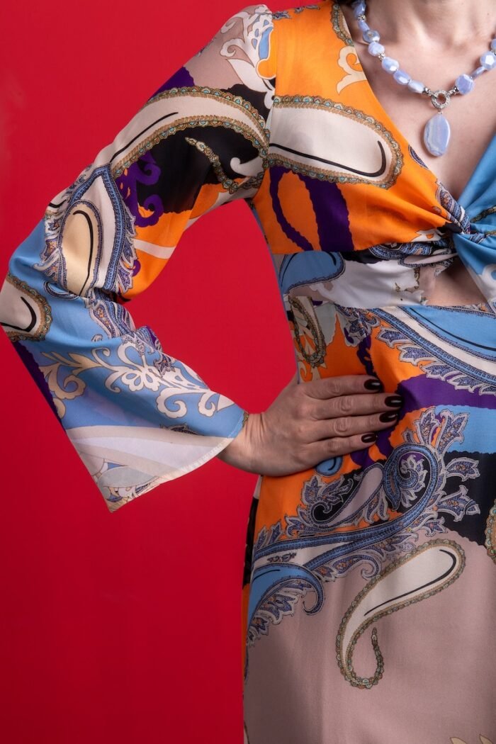 The woman wears a long silk dress with a colorful pattern. She is standing in front of a red background.