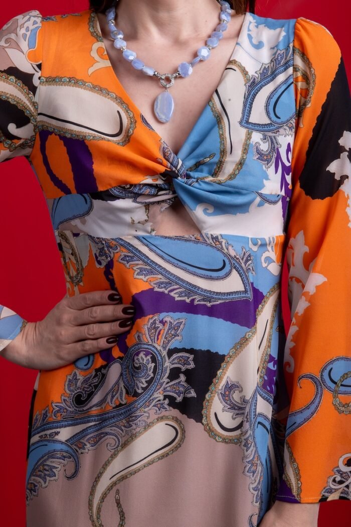 The woman wears a long silk dress with a colorful pattern. She is standing in front of a red background.