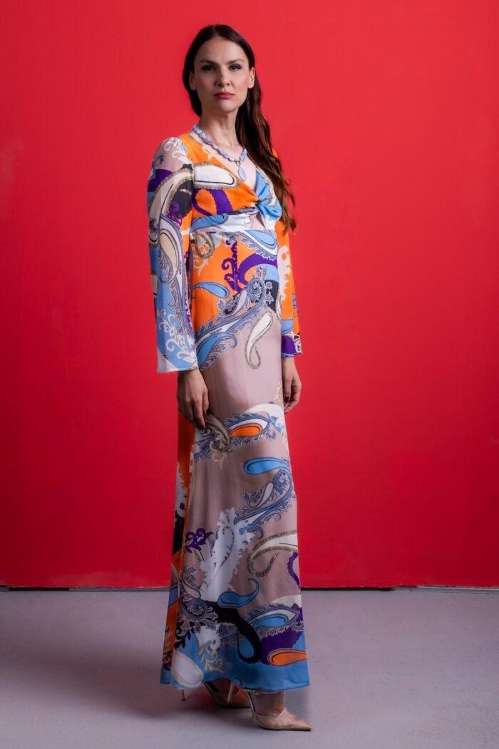 The woman wears a long silk dress with a colorful pattern. She is standing in front of a red background.