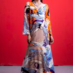 The woman wears a long silk dress with a colorful pattern. He is standing in front of a red background.