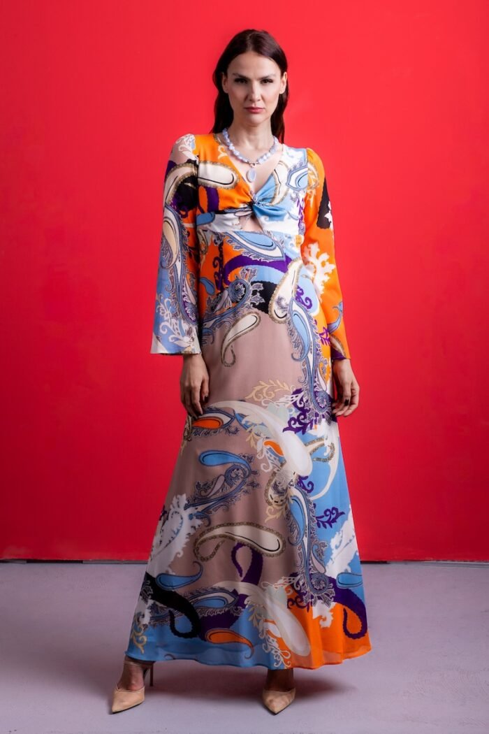 The woman wears a long silk dress with a colorful pattern. He is standing in front of a red background.