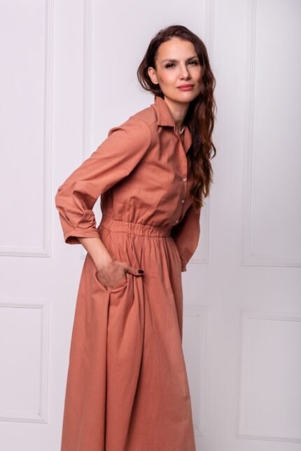 The woman wears a midi dress with a collar. She is standing in front of a white wall.