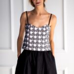 Plava devojka nosi crno-beli top na bretele, crnu suknju i stoji ispred belog zida.