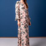 Žena nosi dugu haljinu cvetnog dezena i stoji ispred plave pozadine.