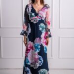 A woman wears a blue and pink patterned dress with long sleeves and stands in front of a white background.